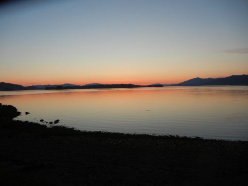 A serene sunset over calm waters, with silhouettes of distant mountains and a colorful sky reflecting on the surface.