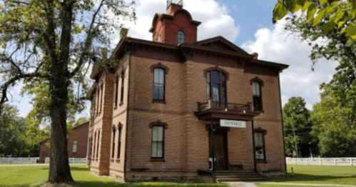Historic brick building with a decorative roof, large windows, and a sign reading "Entrance," surrounded by trees.
