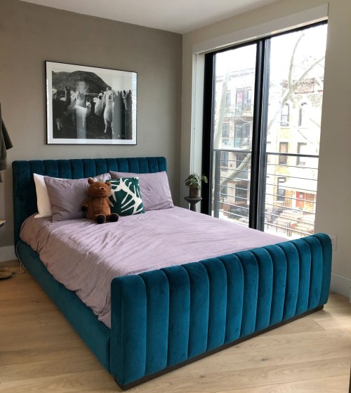 A cozy bedroom featuring a teal bed with a purple blanket, a stuffed animal, and large windows with a city view.