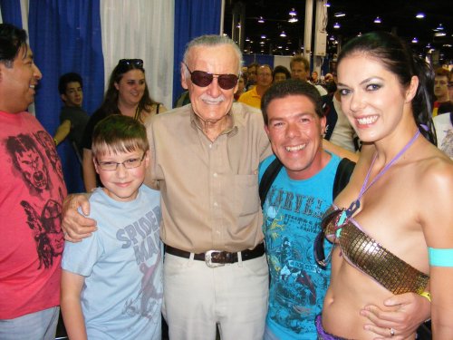 A group photo with a smiling older man in glasses and three fans at a convention, surrounded by a crowd.