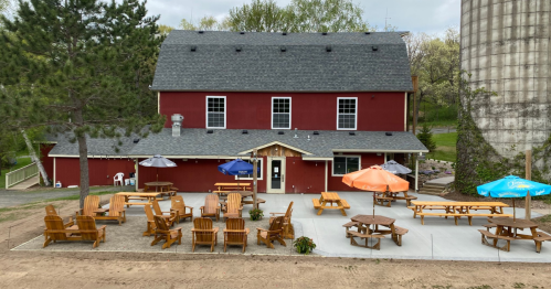 A red barn-style building with outdoor seating, umbrellas, and picnic tables in a grassy area.