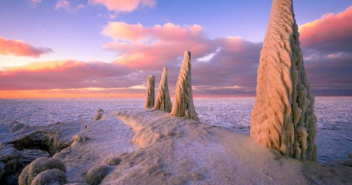 Frozen ice formations rise from a snowy landscape under a colorful sunset sky.