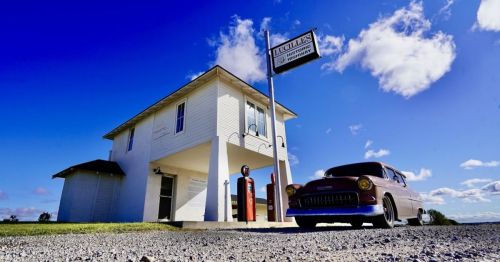 A vintage car parked in front of a white building with a sign, under a bright blue sky with scattered clouds.