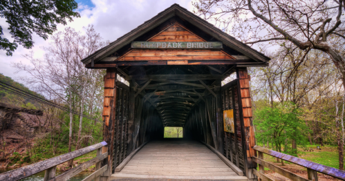A wooden covered bridge with a sign reading "Hayback Bridge," surrounded by trees and a cloudy sky.