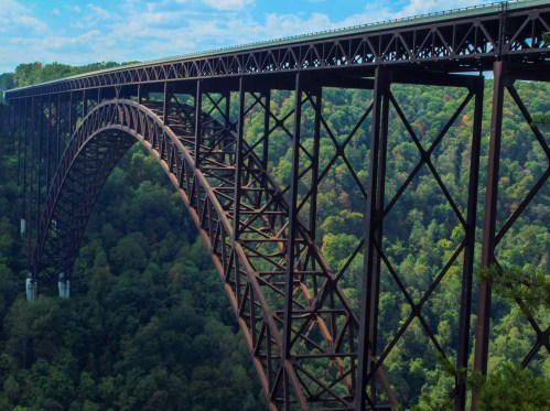 A large steel arch bridge spans a lush green valley under a blue sky with scattered clouds.