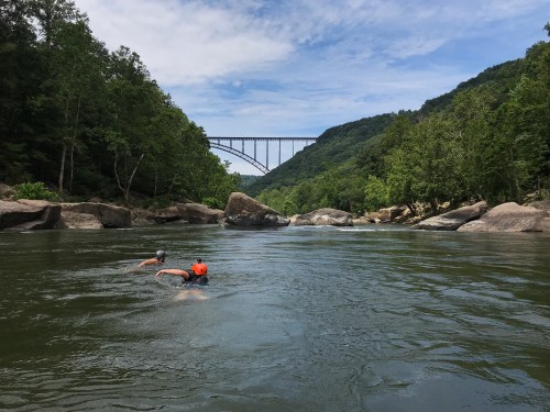 Two swimmers in a river with rocky banks, framed by lush greenery and a bridge in the background under a blue sky.