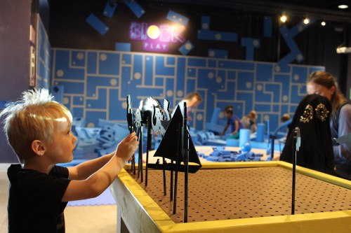 A child plays with black building pieces at a colorful block play area, with other children engaged in the background.