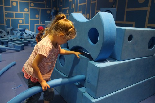 A child plays with large blue foam blocks, exploring and building in a colorful indoor play area.