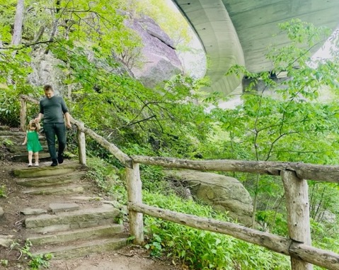 A man and a child walk down a stone path surrounded by greenery, with a bridge overhead.