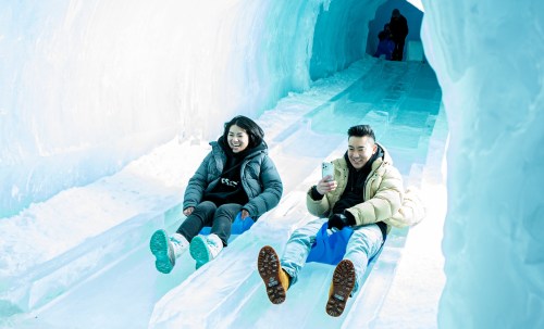 Two people sitting on ice slides, smiling and taking photos in a bright, icy environment.