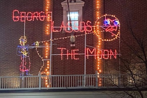 Colorful holiday lights display featuring a figure fishing for the moon, with the text "George Lados" and "The Moon."