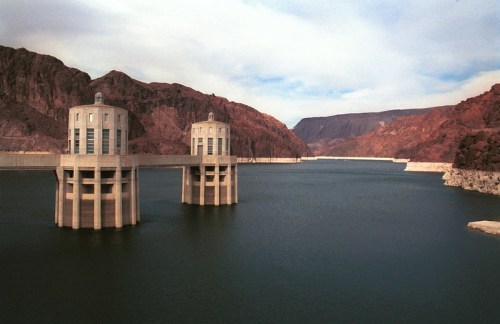 Two large concrete towers stand in a reservoir, surrounded by rocky hills and a cloudy sky.