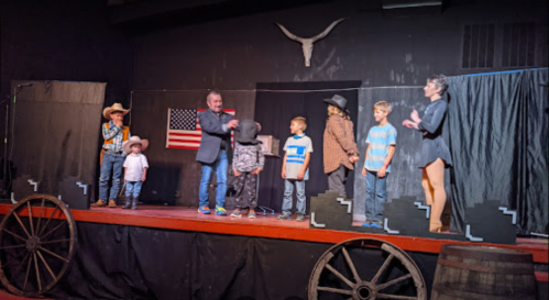 A stage performance featuring children in costumes, a man in a suit, and a woman in a dress, with a Western theme backdrop.