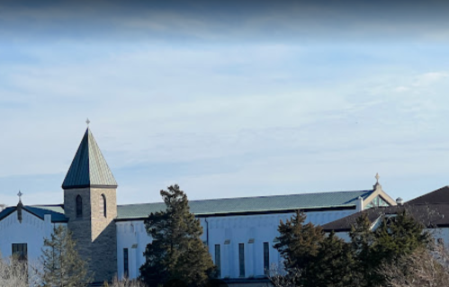 A church building with a tall steeple and a stone facade, surrounded by trees under a clear blue sky.