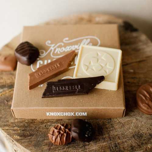 A box of chocolates with two chocolate bars and assorted truffles on a wooden surface.