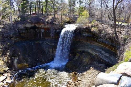 A waterfall cascading into a rocky basin, surrounded by trees and a park setting on a sunny day.