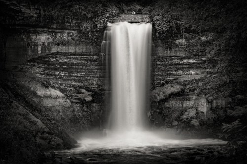 A black and white image of a cascading waterfall surrounded by lush greenery and rocky cliffs.
