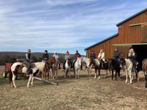 A group of people on horseback in front of a barn, with clear skies and grassy ground.