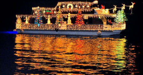 A brightly lit boat decorated with Christmas lights and trees, reflecting on dark water at night.