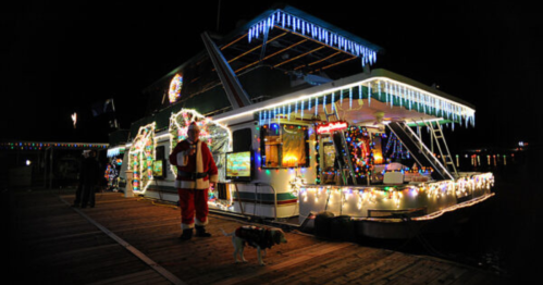 A festive houseboat decorated with lights and icicles, with Santa and a dog standing on the dock at night.