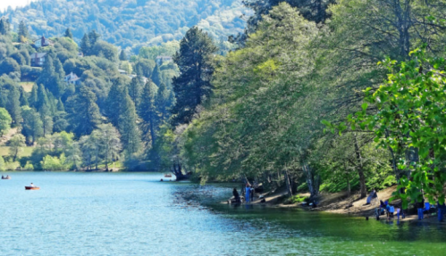 A serene lake surrounded by trees, with people fishing and kayaking on a sunny day. Mountains in the background.