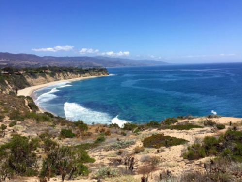 A scenic coastal view with cliffs, sandy beach, and clear blue water under a bright blue sky.