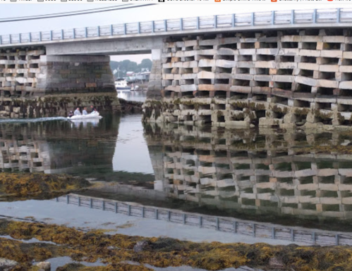 A bridge over calm water, with a boat passing and reflections of the structure and surrounding scenery visible.