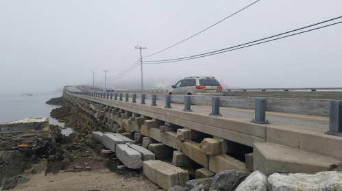 A foggy scene of a road bridge over water, with a car driving and rocky supports visible along the edge.