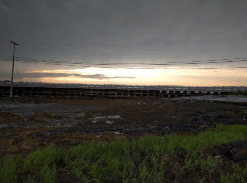 A stormy sky over a bridge with a calm sea, surrounded by wet land and green grass.