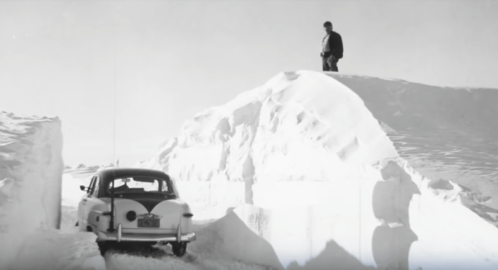 A man stands on a snowbank beside a vintage car navigating through deep snowdrifts.