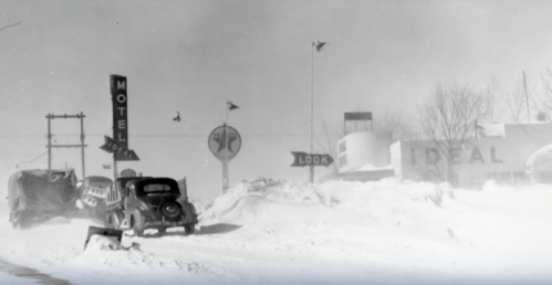 A vintage car drives past a snow-covered roadside with motel signs and heavy snowfall.
