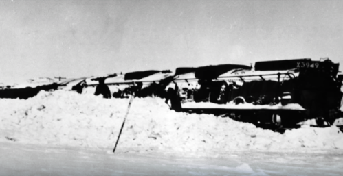 Black and white photo of snow-covered trains, partially buried in deep snow, with people working nearby.