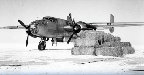 A vintage aircraft parked on snow, with stacks of hay bales nearby.
