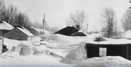 Snow-covered landscape with small buildings and trees, creating a stark winter scene.