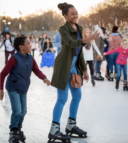 A woman and a boy ice skating, smiling and taking a selfie, with other skaters in the background.