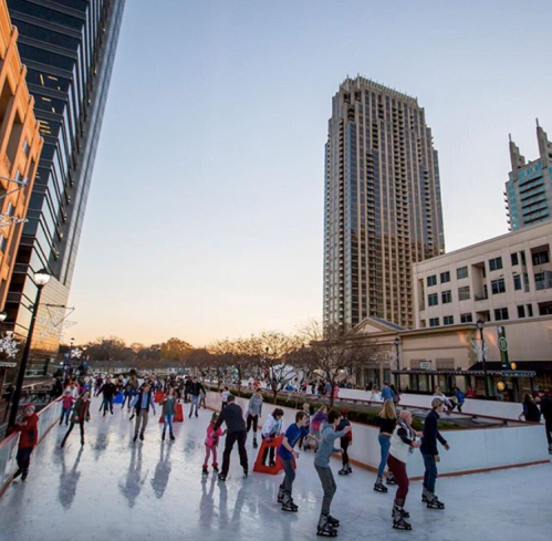 People ice skating on an outdoor rink with city buildings in the background during sunset.