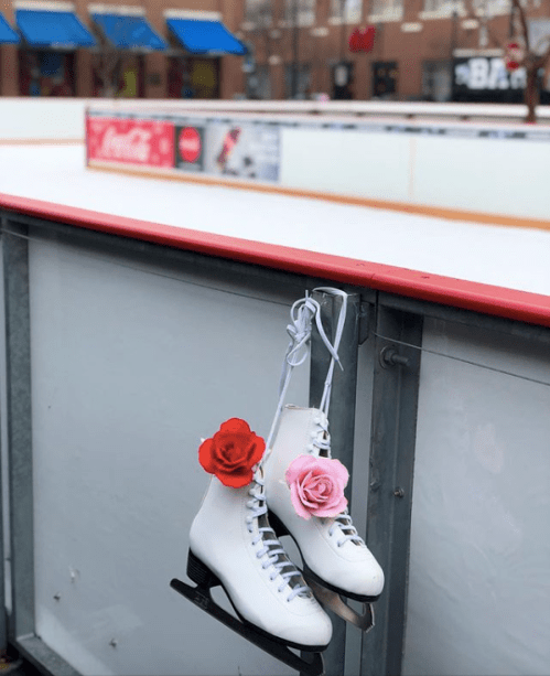 White ice skates with red and pink roses attached, hanging on a rink railing, with an empty ice surface in the background.