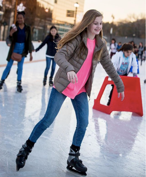 A girl in a pink shirt and jacket skates on ice, smiling, with other skaters in the background.