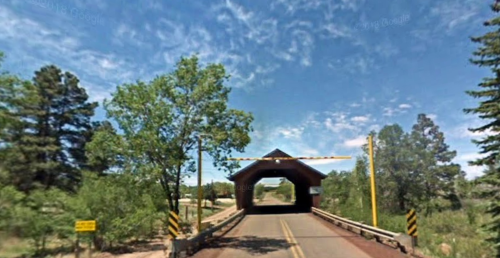 A covered bridge with a wooden structure spans a road, surrounded by trees and a clear blue sky.