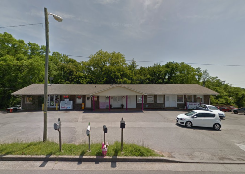 A small commercial building with multiple storefronts, surrounded by greenery and parked cars.