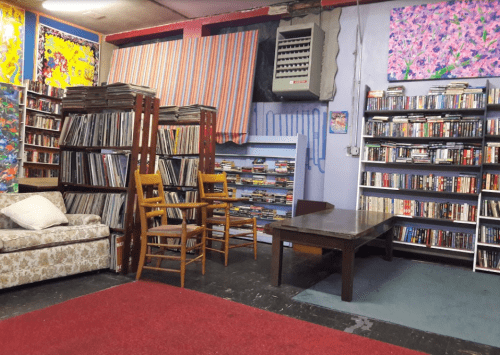 Cozy library space with bookshelves, vinyl records, a table, chairs, and colorful artwork on the walls.