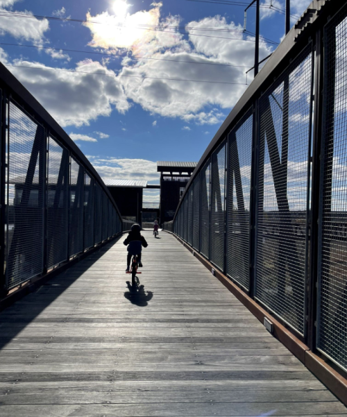 A child rides a bike on a wooden bridge under a bright sky with clouds.