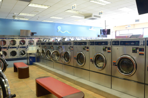 A clean, bright laundromat with rows of washing machines and a red bench for seating.