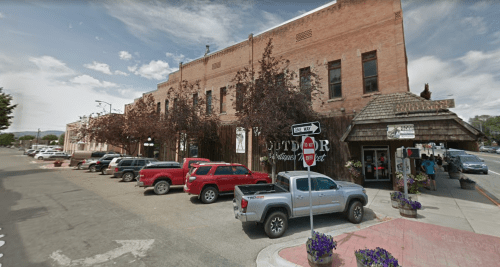 A street view of a small town with brick buildings, parked cars, and shops, under a partly cloudy sky.