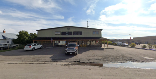 A two-story building with a parking lot, surrounded by vehicles and trees, under a partly cloudy sky.