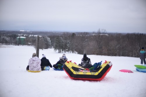 A group of people sitting on sleds at the top of a snowy hill, overlooking a winter landscape.