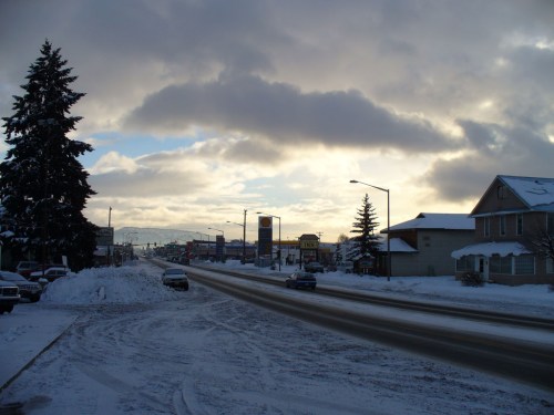 A snowy street scene with cars, trees, and buildings under a cloudy sky at dusk.