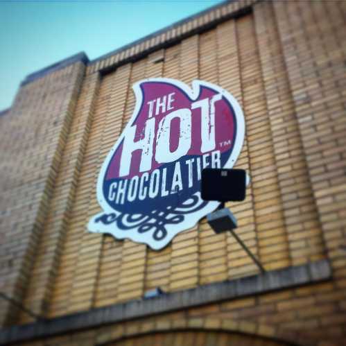 Sign for "The Hot Chocolatier" on a brick wall, featuring a stylized logo in red and white.