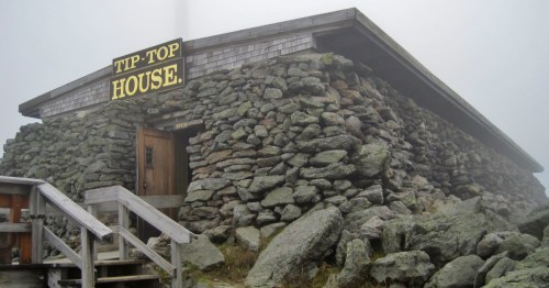 A stone building with a wooden door and a sign reading "TIP-TOP HOUSE," surrounded by rocks and fog.