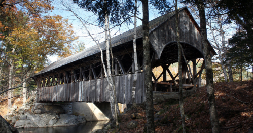 A rustic wooden covered bridge surrounded by trees and autumn foliage, spanning a small stream.